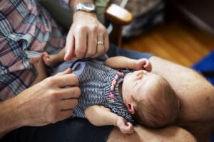 A father holds his newborn baby girl in his lap while sitting in his daughter's nursery at home