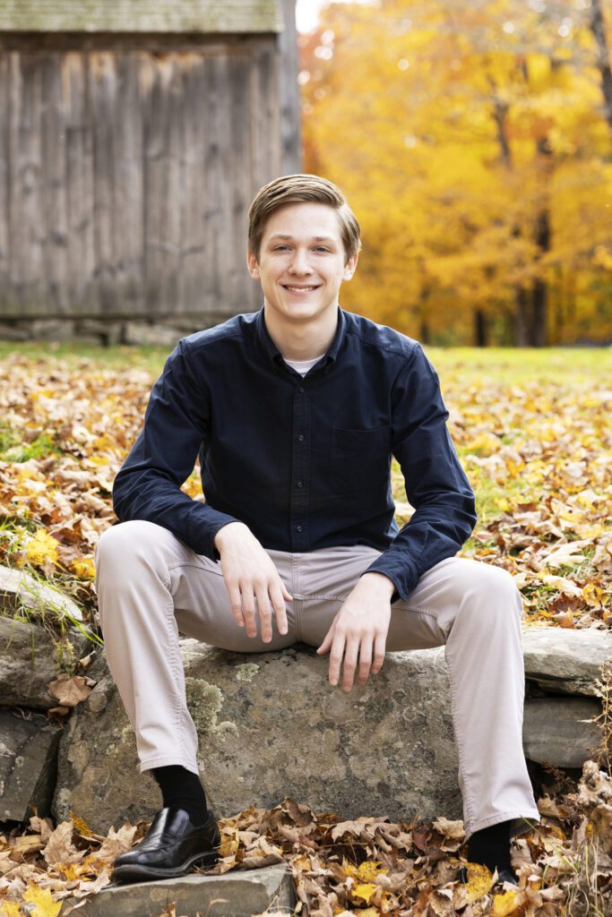 A senior guy in a relaxed portrait with a barn and yellow fall foliage in the background in Groton, Massachusetts